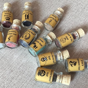 May include: A collection of small glass bottles with cork tops, each filled with a different coloured powder. The bottles are labelled with various names, including "Third Eye Dust", "Fairy of the Scarred Witch Dust", "Money Drawing Dust", "Time is on Me Dust", "Witchbreaker Dust", "Command Dust", "Siren's Dust", "Emerald's Viper Dust", "Victory Dust", "Eternal Separation Dust", and "Trade Dust".