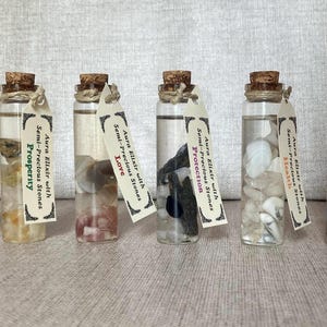 Op de afbeelding: Zes kleine glazen flesjes gevuld met heldere vloeistof en verschillende halfedelstenen. Elk flesje heeft een etiket met de tekst "Aura Elixir with Semi-Precious Stones" en een andere affirmatie: "Open Paths", "Prosperity", "Love", "Protection", "Mental Power" en "Health".
