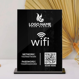 May include: Black acrylic WiFi sign with white text and a QR code. The sign displays the word "wifi" and network information. A gold candlestick and decorative gold geometric shape are on the white surface.