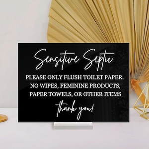 May include: A black sign with white script reading "Sensitive Septic." The sign also says "Please only flush toilet paper. No wipes, feminine products, paper towels, or other items. Thank you!" A gold candlestick and geometric candle holder are also visible.