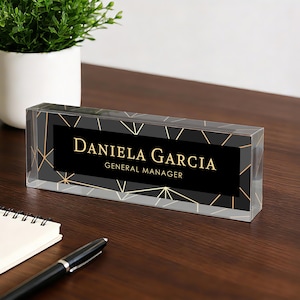 Puede incluir: Una placa de escritorio de acrílico transparente con el nombre "Daniela Garcia" y el título "General Manager" en letras doradas sobre un fondo negro. La placa tiene un patrón geométrico dorado.