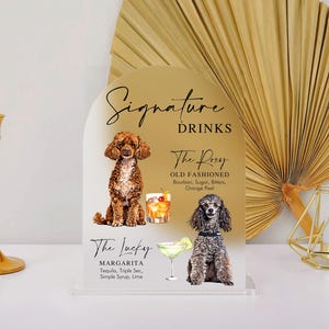 May include: A gold acrylic sign with a rounded top features two illustrations of dogs and text that reads "Signature Drinks". The first drink is "The Roxy Old Fashioned" with the ingredients listed below. The second drink is "The Lucky Margarita" with the ingredients listed below.