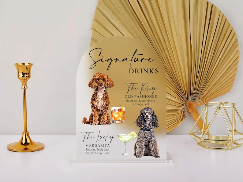 May include: A gold acrylic sign with a rounded top features two illustrations of dogs and text that reads "Signature Drinks". The first drink is "The Roxy Old Fashioned" with the ingredients listed below. The second drink is "The Lucky Margarita" with the ingredients listed below.