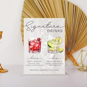 May include: A white sign with the text "Signature Drinks" and two cocktails, one red and one green, with the names "Dirty Shirley" and "Moscow Mule" and their ingredients listed below. The sign also includes the names "Brooke & Jordan" and the date "10.11.20XX".