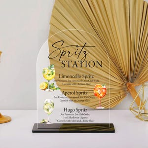 Puede incluir: Un letrero acr&iacute;lico transparente con la inscripci&oacute;n "Spritz STATION" y recetas de c&oacute;cteles. Presenta ilustraciones de spritzes Limoncello, Aperol y Hugo. Un candelabro dorado y un portavelas geom&eacute;trico est&aacute;n a ambos lados. El fondo incluye una hoja de palma seca.