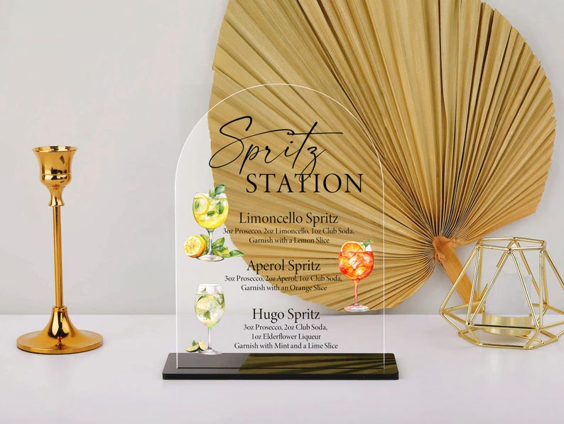 Puede incluir: Un letrero acr&iacute;lico transparente con la inscripci&oacute;n "Spritz STATION" y recetas de c&oacute;cteles. Presenta ilustraciones de spritzes Limoncello, Aperol y Hugo. Un candelabro dorado y un portavelas geom&eacute;trico est&aacute;n a ambos lados. El fondo incluye una hoja de palma seca.