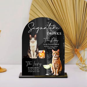 May include: Black sign with white text that reads "Signature Drinks". The sign features two cocktails, "The Roxy Old Fashioned" and "The Lucky Margarita", with illustrations of cats.