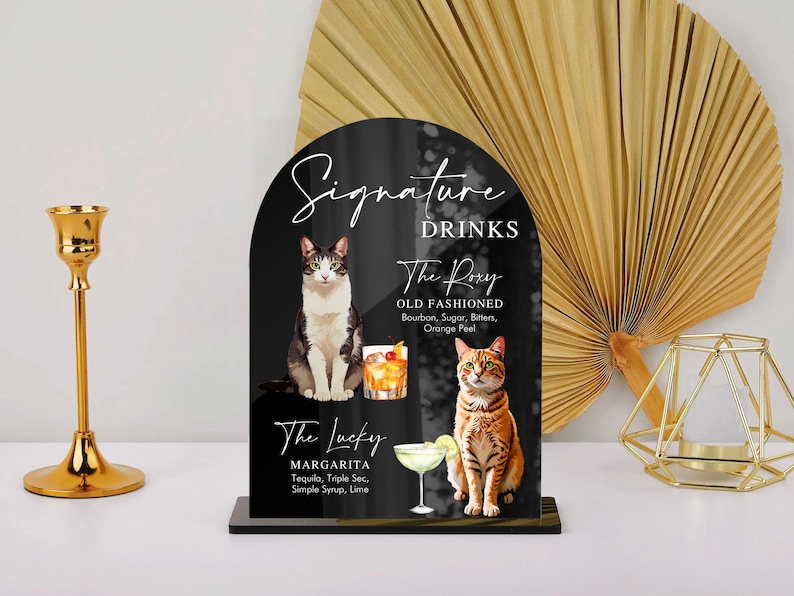 May include: Black sign with white text that reads "Signature Drinks". The sign features two cocktails, "The Roxy Old Fashioned" and "The Lucky Margarita", with illustrations of cats.