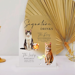 May include: A white acrylic sign with a gold arch design. The sign features two cats and text that reads "Signature Drinks", "The Pory Old Fashioned", "Bourbon, Sugar, Bitters, Orange Peel", "The Lucky Margarita", "Tequila, Triple Sec, Simple Syrup, Lime".