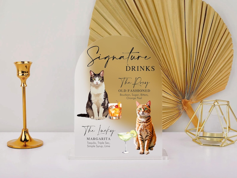 May include: A white acrylic sign with a gold arch design. The sign features two cats and text that reads "Signature Drinks", "The Pory Old Fashioned", "Bourbon, Sugar, Bitters, Orange Peel", "The Lucky Margarita", "Tequila, Triple Sec, Simple Syrup, Lime".