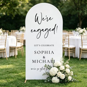 May include: A white arched sign with black text reading We're engaged! LET'S CELEBRATE SOPHIA & MICHAEL we're so glad you're here. The sign is decorated with a cluster of white hydrangea and rose flowers with green leaves at the bottom right corner.