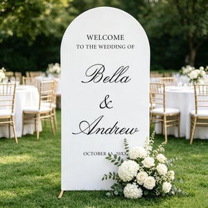 Op de afbeelding: Een wit boogvormig welkomstbord voor een bruiloft met zwarte tekst WELCOME TO THE WEDDING OF Bella & Andrew OCTOBER 13, 20XX. Het bord is aan de onderkant versierd met een boeket witte hortensia's en rozen met groene bladeren.