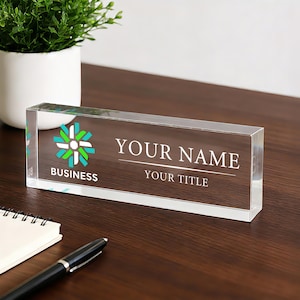Puede incluir: Una placa de escritorio de acrílico transparente con el texto "YOUR NAME" y "YOUR TITLE" en fuente blanca. La placa también presenta un logotipo y la palabra "BUSINESS". Una pequeña planta en maceta, un cuaderno y un bolígrafo también están en el escritorio.