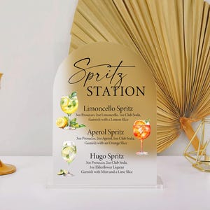Puede incluir: Un letrero dorado de "Spritz Station" con recetas de c&oacute;cteles, incluyendo Limoncello, Aperol y Hugo Spritz. El letrero est&aacute; sobre una base de acr&iacute;lico transparente. Tambi&eacute;n se ven un candelabro dorado, una decoraci&oacute;n geom&eacute;trica dorada y un abanico de hojas de palma secas.