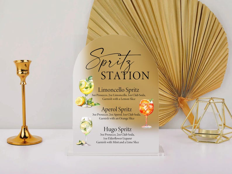 Puede incluir: Un letrero dorado de "Spritz Station" con recetas de c&oacute;cteles, incluyendo Limoncello, Aperol y Hugo Spritz. El letrero est&aacute; sobre una base de acr&iacute;lico transparente. Tambi&eacute;n se ven un candelabro dorado, una decoraci&oacute;n geom&eacute;trica dorada y un abanico de hojas de palma secas.