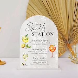 Puede incluir: Un letrero de acr&iacute;lico transparente con una parte superior arqueada, etiquetado como "Spritz STATION". Presenta ilustraciones y recetas de c&oacute;cteles Limoncello, Aperol y Hugo Spritz. Un candelabro dorado y una decoraci&oacute;n geom&eacute;trica a cada lado.