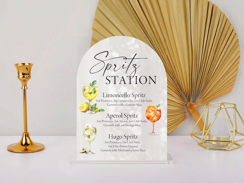 Puede incluir: Un letrero de acr&iacute;lico transparente con una parte superior arqueada, etiquetado como "Spritz STATION". Presenta ilustraciones y recetas de c&oacute;cteles Limoncello, Aperol y Hugo Spritz. Un candelabro dorado y una decoraci&oacute;n geom&eacute;trica a cada lado.