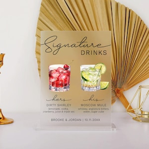May include: A white acrylic sign with black text that reads "Signature Drinks". The sign features two watercolor illustrations of cocktails, one red and one green, with the text "hers" and "his" below each. The sign also includes the names of the cocktails, "Dirty Shirley" and "Moscow Mule", and their ingredients. The bottom of the sign reads "Brooke & Jordan | 10.11.20XX".