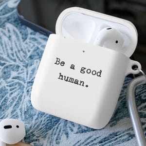 Puede incluir: Funda blanca para AirPods con el texto "Be a good human." La funda está abierta, revelando auriculares blancos. Un mosquetón plateado está sujeto a la funda. El fondo incluye un smartphone y una tela estampada en azul y blanco.
