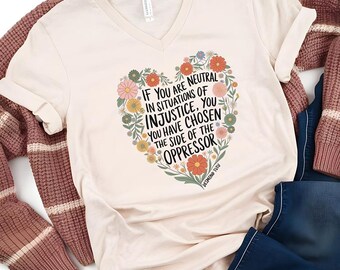 May include: A cream-colored V-neck t-shirt with a floral heart design and the text "IF YOU ARE NEUTRAL IN SITUATIONS OF INJUSTICE, YOU HAVE CHOSEN THE SIDE OF THE OPPRESSOR." The shirt is paired with a brown sweater and blue jeans.