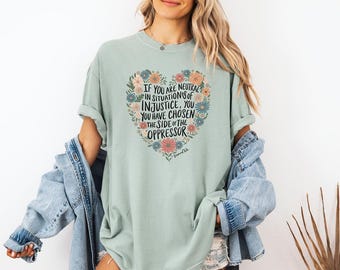 May include: A light blue t-shirt with a floral heart design and the text "IF YOU ARE NEUTRAL IN SITUATIONS OF INJUSTICE, YOU HAVE CHOSEN THE SIDE OF THE OPPRESSOR".