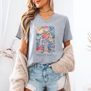 K&ouml;nnte beinhalten: Graues V-Ausschnitt-T-Shirt mit einem Blumendesign und dem Text "Resist Hate" auf der Vorderseite.