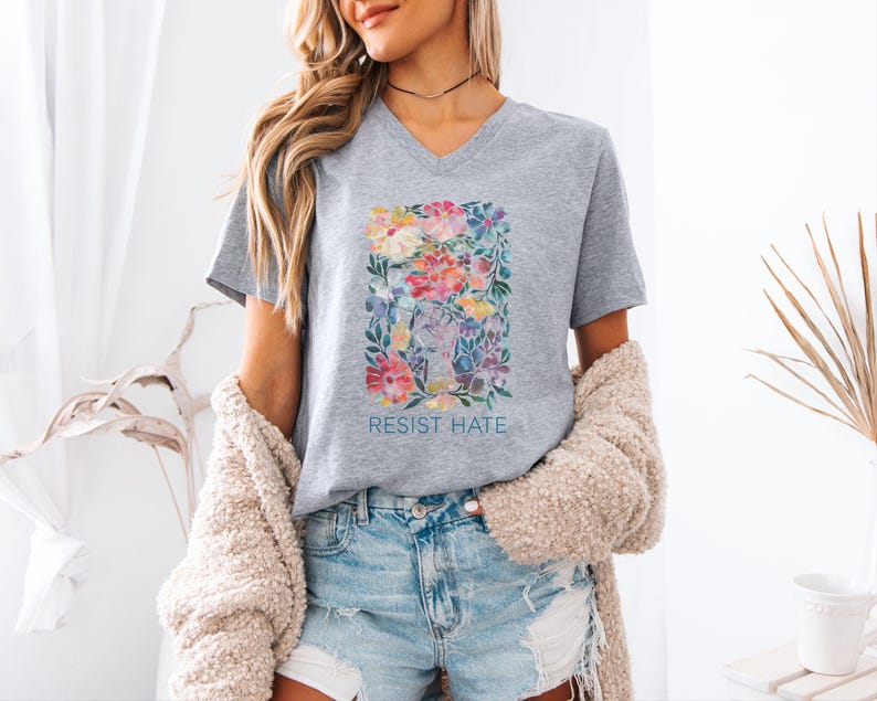K&ouml;nnte beinhalten: Graues V-Ausschnitt-T-Shirt mit einem Blumendesign und dem Text "Resist Hate" auf der Vorderseite.