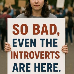 May include: A person wearing a black beanie and a grey shirt holds a white sign with the text "SO BAD, EVEN THE INTROVERTS ARE HERE." in red and blue lettering.