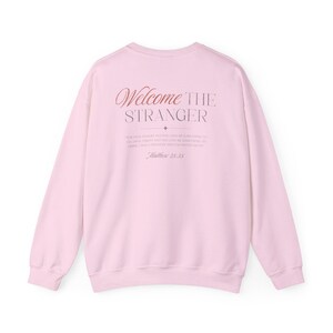 May include: A light pink sweatshirt with the words "Welcome the Stranger" in a decorative font on the back. Below the text is a quote from Matthew 25:35.