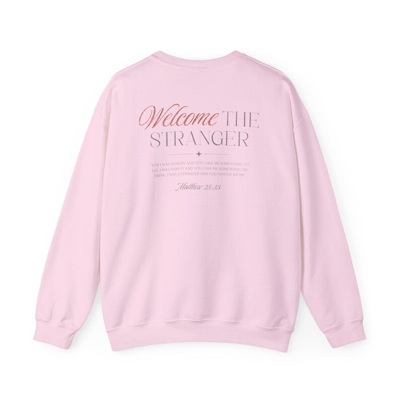 May include: A light pink sweatshirt with the words "Welcome the Stranger" in a decorative font on the back. Below the text is a quote from Matthew 25:35.