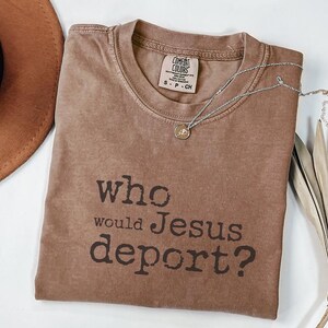 K&ouml;nnte beinhalten: Ein braunes T-Shirt mit dem Text "who would Jesus deport?" in dunkelbraunen Buchstaben. Eine silberne Halskette mit einem kleinen Anh&auml;nger befindet sich auf dem Shirt. Ein brauner Hut und getrocknete Bl&auml;tter sind ebenfalls im Bild.