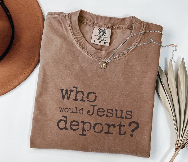 K&ouml;nnte beinhalten: Ein braunes T-Shirt mit dem Text "who would Jesus deport?" in dunkelbraunen Buchstaben. Eine silberne Halskette mit einem kleinen Anh&auml;nger befindet sich auf dem Shirt. Ein brauner Hut und getrocknete Bl&auml;tter sind ebenfalls im Bild.