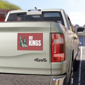 May include: A rectangular red bumper sticker with the text "NO KINGS" in white, and an illustration of the Statue of Liberty. The sticker is affixed to the back of a light green pickup truck.