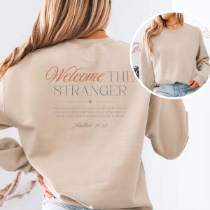 May include: Beige sweatshirt with the text "Welcome the Stranger" in a script font on the back. Below the text is a quote from Matthew 25:35. The sweatshirt has long sleeves and a crew neck.