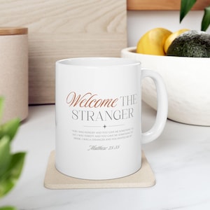 May include: White ceramic mug with the words "Welcome the Stranger" in a decorative font. Below the text is a quote from Matthew 25:35. The mug sits on a beige coaster.