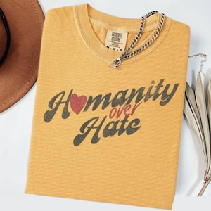 May include: Mustard yellow t-shirt with the words "Humanity over Hate" in a retro font. The word "Humanity" has a red heart in place of the letter "o". A silver chain necklace rests on the shirt.