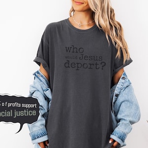 K&ouml;nnte beinhalten: Dunkelgraues T-Shirt mit dem schwarzen Text "who would Jesus deport?". Das Shirt wird mit einer hellblauen Jeansjacke getragen. Eine Sprechblase zeigt "10% of profits support social justice."