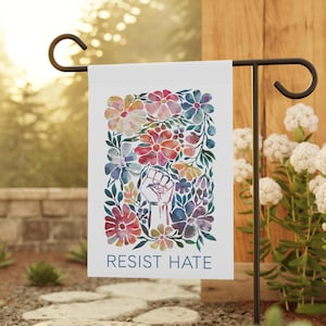 A white garden flag with a watercolor design of colorful flowers and a raised fist. The text 'RESIST HATE' is printed in black at the bottom of the flag.