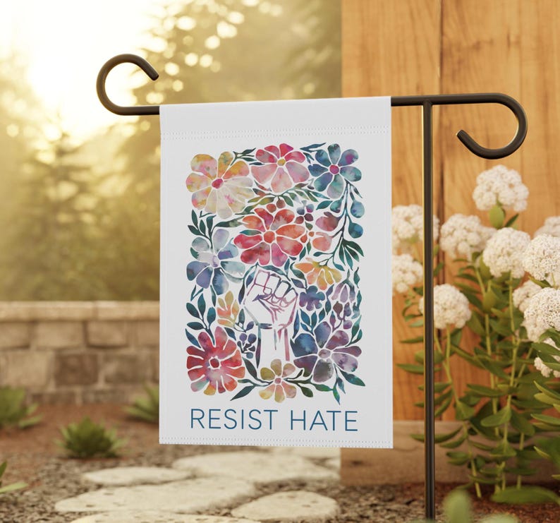 A white garden flag with a watercolor design of colorful flowers and a raised fist. The text 'RESIST HATE' is printed in black at the bottom of the flag.