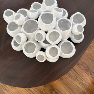 Pode incluir: Uma escultura de cerâmica branca com múltiplas formas arredondadas e abertas. A escultura tem uma superfície texturizada e está disposta em um padrão agrupado. O interior de cada forma é de cor cinza claro. A escultura é exibida em uma superfície marrom escuro.