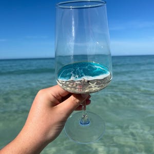 Könnte beinhalten: Ein klares Weinglas mit einer Miniatur-Strandszene im Inneren, gehalten vor dem Meer und einem blauen Himmel. Das Glas enthält Sand, Muscheln und ein Harz-Ozean-Design mit weißen Wellen und türkisfarbenem Wasser.