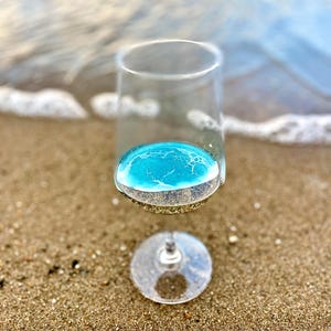 May include: A clear glass wine glass filled with a blue liquid with white foam on top, sitting on a sandy beach.