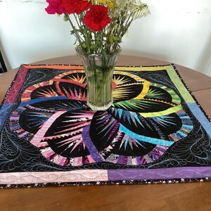 May include: A colourful quilted table runner with a black background and a floral design. The runner has a pink, purple, blue, green and yellow border.