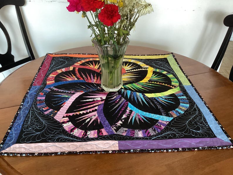 May include: A colourful quilted table runner with a black background and a floral design. The runner has a pink, purple, blue, green and yellow border.