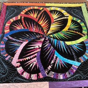 May include: A colourful quilt with a black background and a swirling design of geometric shapes. The quilt is made of various fabrics in shades of red, orange, yellow, green, blue, purple, and pink.