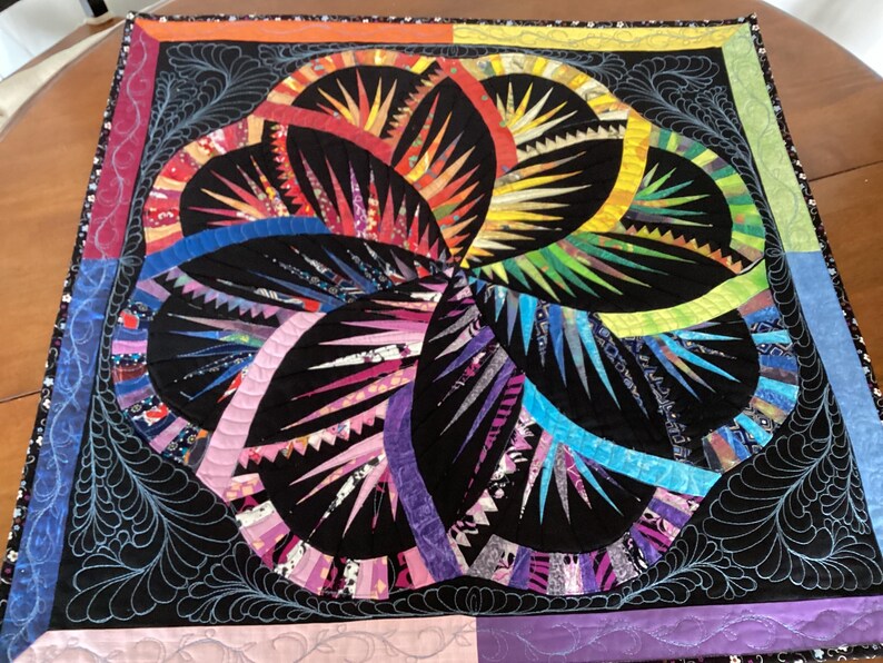 May include: A colourful quilt with a black background and a swirling design of geometric shapes. The quilt is made of various fabrics in shades of red, orange, yellow, green, blue, purple, and pink.