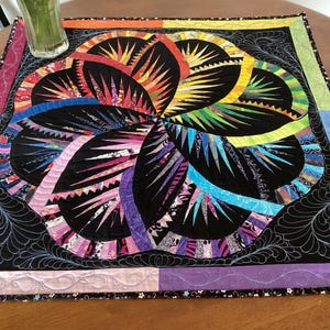 May include: A colourful quilt with a black background and a large, multi-coloured flower design. The flower is made up of many different fabric scraps, and the quilt is edged with a black and white trim.