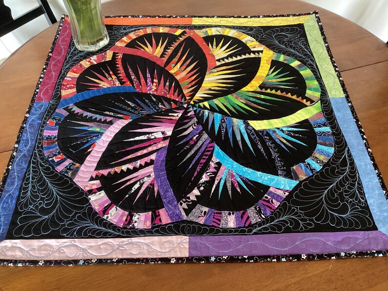 May include: A colourful quilt with a black background and a large, multi-coloured flower design. The flower is made up of many different fabric scraps, and the quilt is edged with a black and white trim.