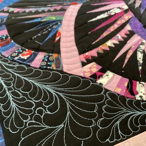 May include: Close-up of a black fabric with a blue feather-like quilting design. The fabric is part of a larger quilt with a colourful patchwork design.