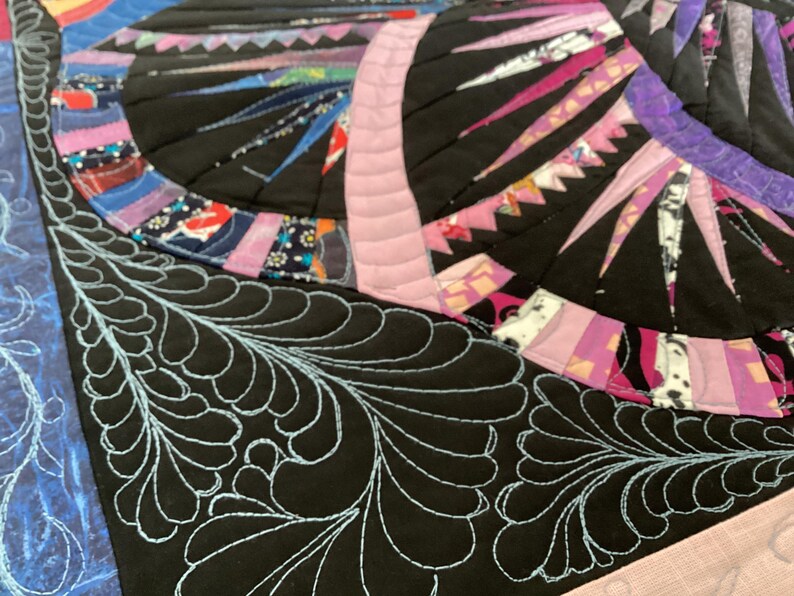 May include: Close-up of a black fabric with a blue feather-like quilting design. The fabric is part of a larger quilt with a colourful patchwork design.